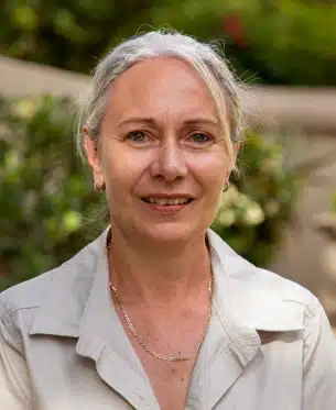 A portrait of an older woman with short gray hair, smiling subtly at the camera. She is wearing a light beige shirt and a delicate necklace depicting African Safaris. The background is softly blurred with green
