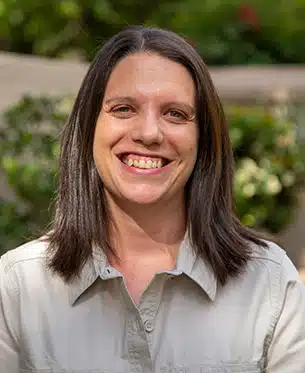 A portrait of a smiling woman with straight dark brown hair. She wears a light gray shirt and stands outdoors with green blurred foliage in the background from her recent African safaris. She appears cheerful and approach