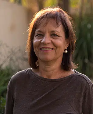 A smiling middle-aged woman with short brown hair and earrings, wearing a brown top. She is photographed outdoors with soft-focused greenery in the background, reminiscent of an award-winning African safari setting.