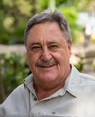 A portrait of a smiling elderly man with a mustache, wearing a white shirt. He has short gray hair, wrinkles, and a warm expression, set against a blurred green foliage background reminiscent of an