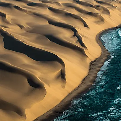 Aerial view of a coastline in Namibia where towering sand dunes meet the deep blue ocean, creating a dramatic natural contrast between the golden sands and the water.