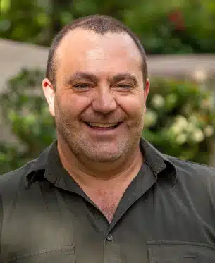An award-winning middle-aged man with a hearty smile and short dark hair, wearing a dark green shirt, stands outdoors with green foliage softly blurred in the background.