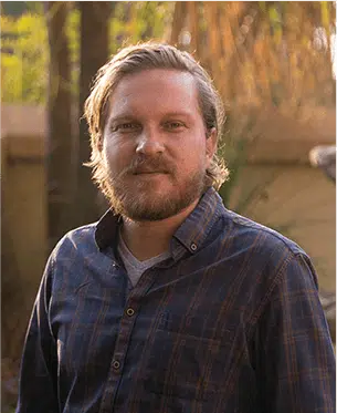 A portrait of a smiling man with a beard and mustache. He wears a blue plaid shirt and stands outdoors with trees softly blurred in the background, conveying a warm, golden hour light characteristic of