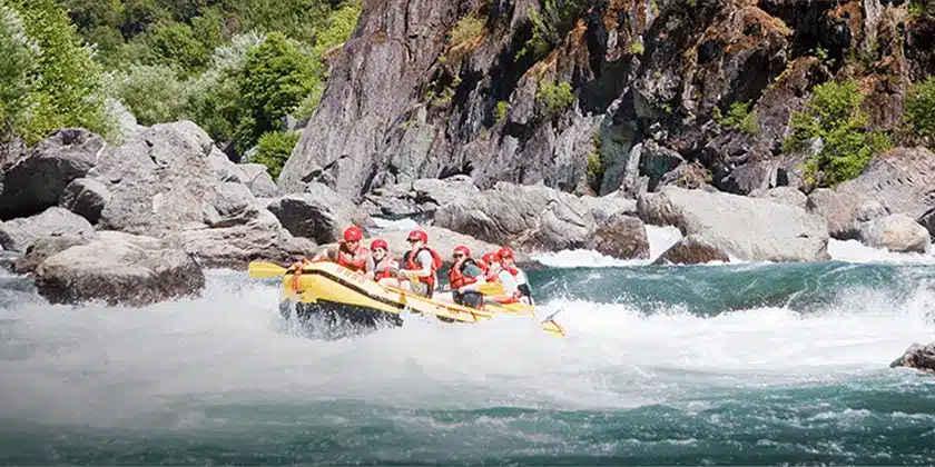 A group of six people in red life jackets is white water rafting on a turbulent river, navigating through rocky terrain, amidst lush greenery and jagged cliffs, experiencing the thrill of adventure travel.