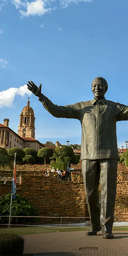 A tall statue of Nelson Mandela with an outstretched arm, positioned in a sunny plaza. In the background, green terraced gardens and an ornate building with a tower under a clear blue sky