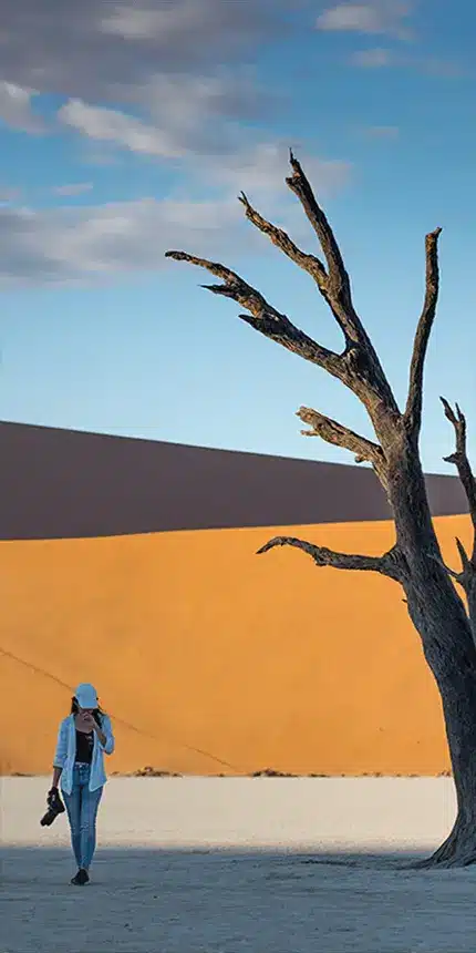 A woman walks near a stark, leafless tree against a backdrop of a smooth sand dune and a clear blue sky. She is wearing a hat, sunglasses, and carries a camera, emphasizing a