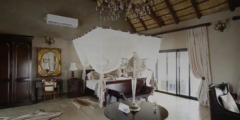 An elegant, vintage-style room with a wooden floor and high ceiling, featuring a large draped canopy bed, antique furniture, ornate chandelier, and large glass doors leading to a luxury safari experience outside