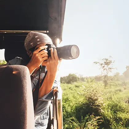 A photographer wearing a cap and a denim shirt leans out of a luxury safari vehicle, using a long camera lens to capture photos of a sunlit landscape with lush greenery.