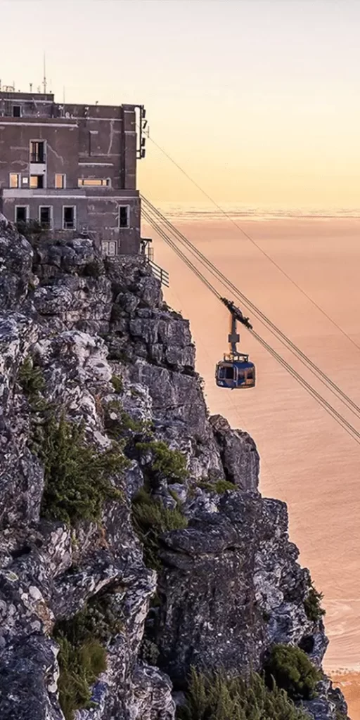 A cable car ascends a steep cliff towards a building at sunset, surrounded by a vast, soft-hued sky and scattered clouds, highlighting the rugged textures of the rocky cliff's surface. This scene