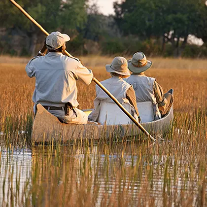 Three individuals in a canoe on a river at sunset, embarking on a serene travel experience. Two are in hats, and one, standing, uses a pole to navigate. Tall grass lines the river