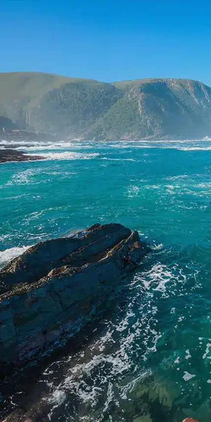 Turquoise ocean waves crash against jagged rocks with lush green mountains under a clear blue sky in the background. A serene coastal scene exuding vibrant natural colors and dynamic water movement, offering a luxury experience