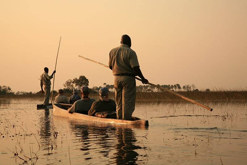 Two people guide canoes through a calm, reedy waterway at sunset, silhouetted against a golden sky. One stands navigating with a pole, and the other, seated, paddles