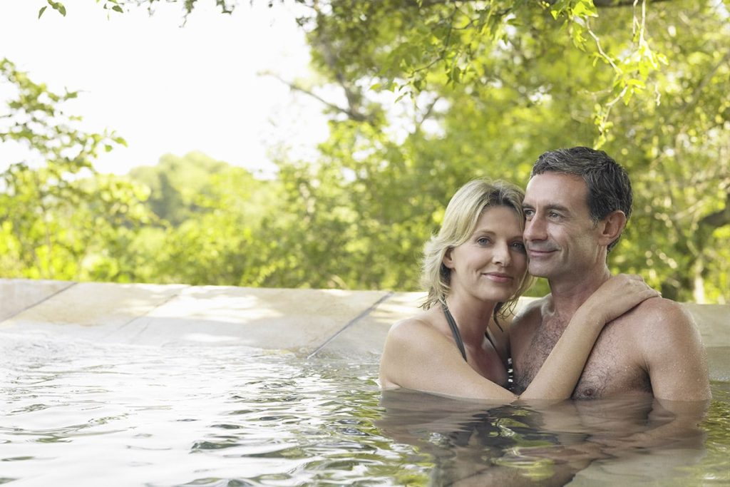 A middle-aged Caucasian couple embraces in a swimming pool surrounded by lush greenery, smiling contentedly during their idyllic honeymoon. The woman rests her head on the man's shoulder, both partially submerged