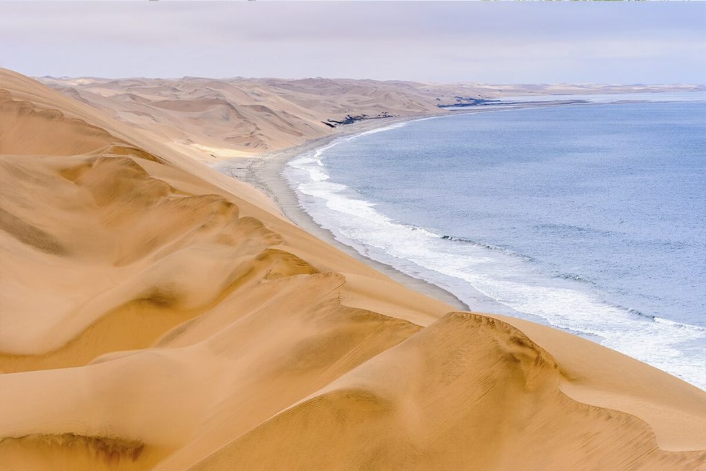 A panoramic view of the Namib Desert coastline near Swakopmund showcasing a series of large golden sand dunes cascading down to a tranquil blue ocean. Waves gently lap against the shoreline, contrasting