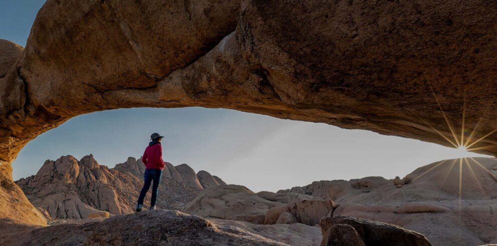 A person in a red jacket and dark hat stands on a rocky terrain, beneath a massive natural rock arch. The sun is low, casting a warm light and starburst effect to the right, illuminating the rugged landscape reminiscent of Namibia's safari regions with boulders and mountains in the background under a clear sky.