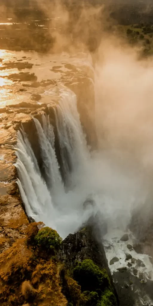 Aerial view of a majestic waterfall in Zimbabwe, with voluminous water cascading down a steep cliff amidst lush greenery, as mist rises and the golden light of sunset reflects off the water and surrounding