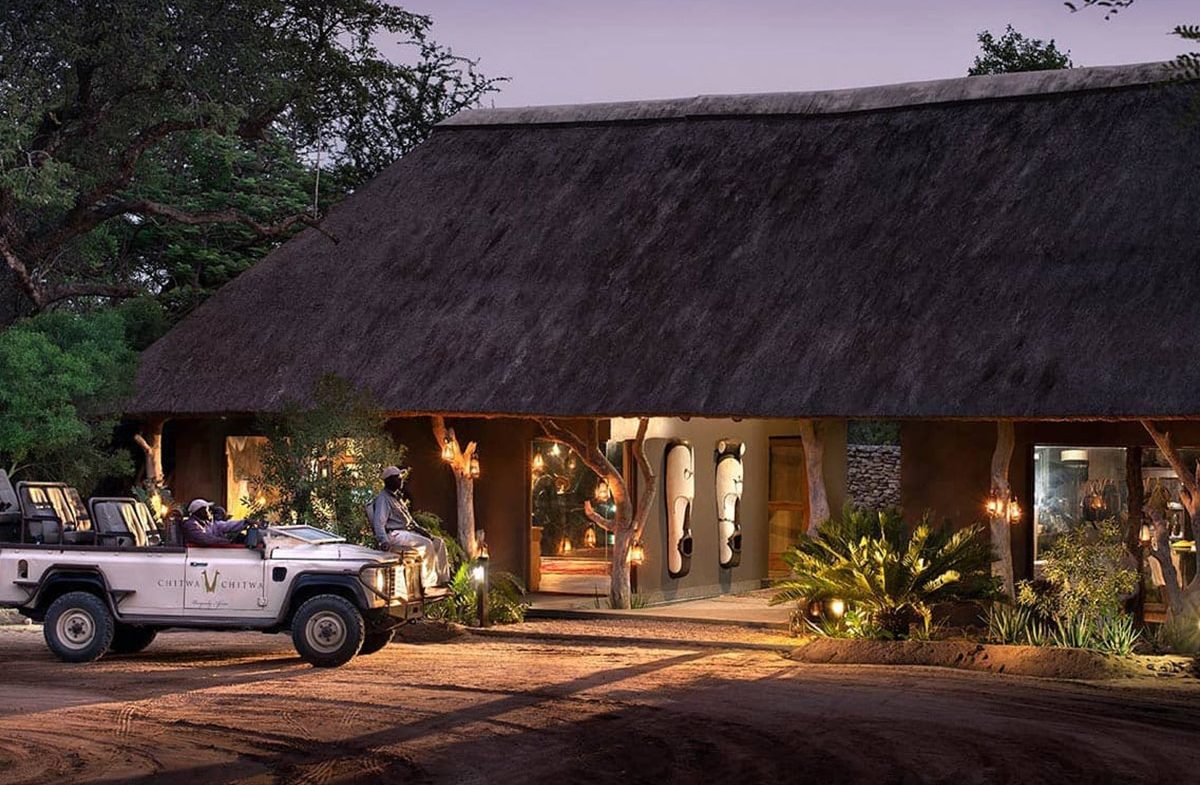 Two people in a safari vehicle parked outside a luxury lodge illuminated by warm lights at dusk, with lush plants surrounding the entrance.