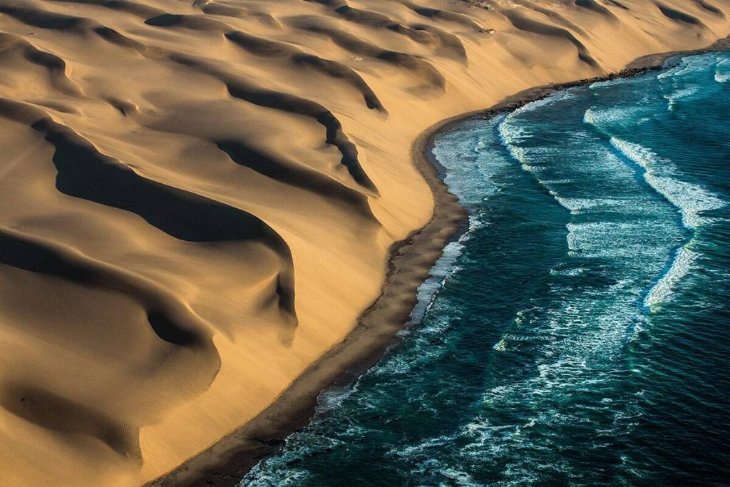 Aerial view of a coastal desert landscape in Namibia where golden sand dunes meet the blue ocean, creating a striking contrast between the soft, rippled dune patterns and the wavy, fo
