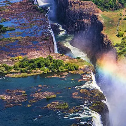 Aerial view of a majestic waterfall with a vibrant rainbow near its base. Lush greenery surrounds the top and banks of the river, and a series of small, rocky islands dot the water below