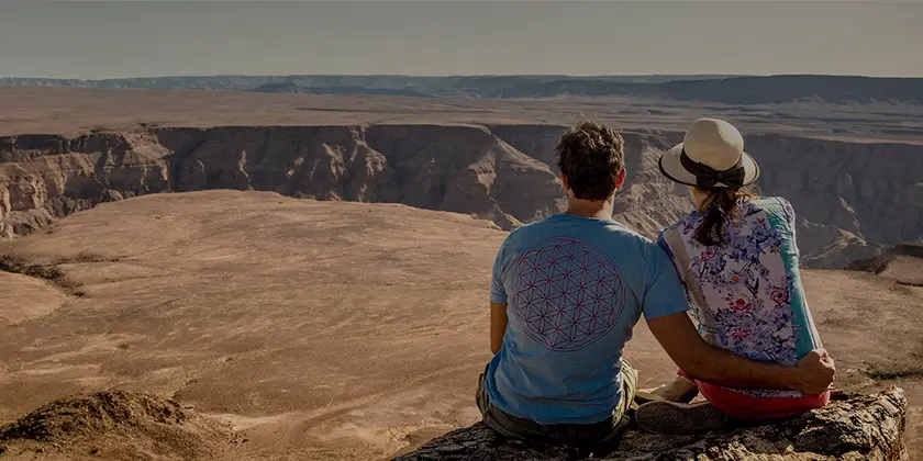 A couple sits close together on a rocky ledge overlooking a vast canyon under a clear blue sky. In this luxury travel experience, the man wears a blue shirt with a red design, and the woman sports