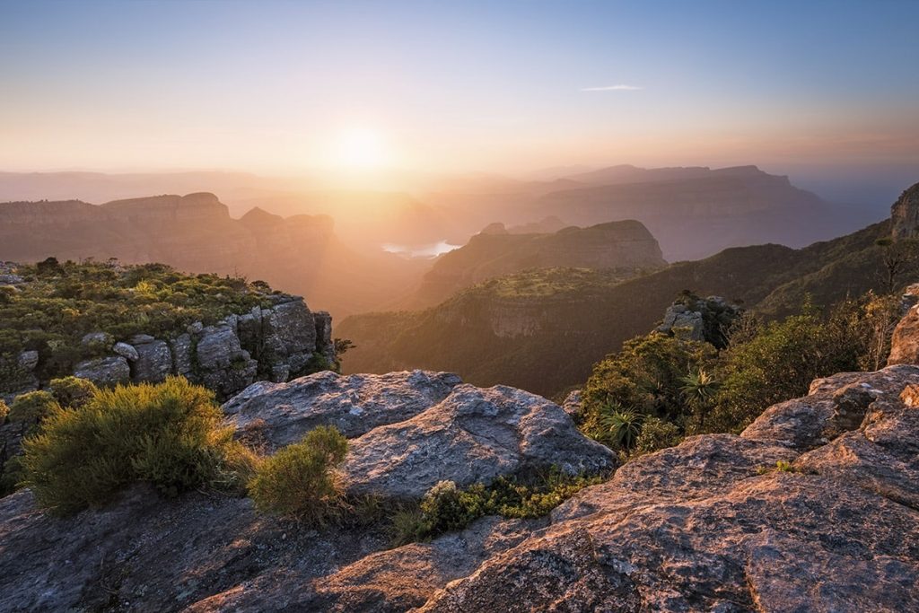 A scenic vista at sunrise showcases a rugged canyon with jagged cliffs and dense vegetation in South Africa. The sun, low in the sky, illuminates the valley and touches the tips of the cliffs,