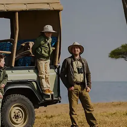 Two individuals dressed in safari gear stand next to an open safari vehicle by a scenic lake. One is stepping out of the vehicle, while the other stands beside it, both ready for an exhilarating travel